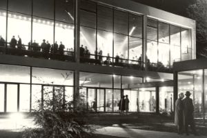 Ein Haus mit großer Glasfassade, innen hell erleuchtet, das obere Stockwerk voller Menschen.