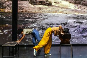 Zwei Performer*innen sind über einen Stuhl gebeugt. Sie stützen sich auf diese und scheinen sie aus dem Bild schieben zu wollen. Im Hintergrund zeigt eine Videoprojektion Wasser, dass schnell über flache Kaskaden fließt.  