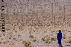 Die Collage zeigt ein Foto von Ada Mukhína in einer Rückenansicht, wie sie in die Weite einer Wüste blickt. Über die Aufnahme ist ein alter Stadtplan von Berlin-Wilmersdorf gelegt.