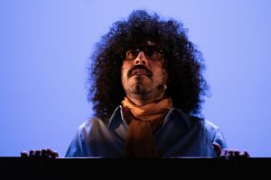 Die Farbfotografie zeigt eine Nahaufnahme von einer Person vor einem blauleuchtenden Hintergrund. Sie hat lange schwarze Locken, trägt eine Brille auf schaut nach oben in den Bühnenhimmel.  