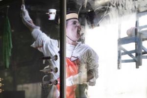 Idiota in der Glasvitrine hat eine rote Schürze und weißen Handschuhe an und presst den geöffneten Mund und die Nase an der Scheibe platt. Auf Kopfhöhe ist ein Becher befestigt. Darunter drei mit Farbe aufgezogene Spritzen.