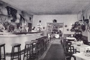 Eine noch menschenleere, aufgeräumte Bar mit vielen Pflanzen und Wanddekoration. Im vorderen Teil links: ein Tresen mit Barhockern. Rechts: Tische mit Gedecken. Ein Durchbruch lässt im nächsten Raum Instrumente erkennen, die auf die Band warten.