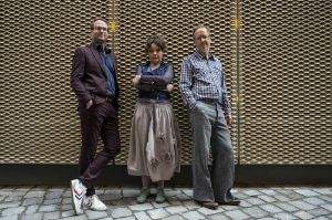 The trio "Vesna" (Spring) a project by Mariana Sadovska stands in front of a facade with strong relief. She is wearing a leather vest and skirt and holds her arms crossed. To her right and left, Christian Thomé and Markus Braun lean against the wall.
