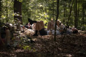 Several people are lying with their backs on the forest floor, which is covered in leaves. They are wearing headphones.