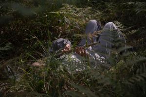 Eine Person in einem grauen Overall liegt mit dem Rücken auf dem Waldboden und spielt Querflöte. Im grünen Dickicht ist ihr Gesicht nicht zu sehen.