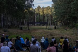 Eine Gruppe Menschen sitzt auf einer Wiese. Sie schauen auf einen länglichen LED-Bildschirm, der in der Landschaft steht. Auf dem Bildschirm leuchten die Buchstaben „Ich sehe euch“.