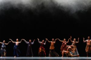 A dancer grabs his throat with a deep lunge while other dancers form a chain behind him.