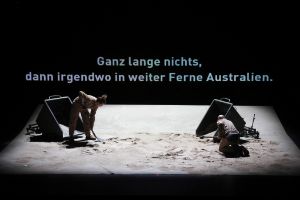 Five people are in a kind of beach scene, above them is written as a projection “Was ist eine Erinnerungsnotiz wert, wenn man nicht mehr weiß, dass man nach ihr suchen könnte (What is a memory note worth if you don’t know you could look for it any more)?”