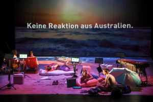 Two people shovel sand from the stage, above them is written as a projection on a screen “Ganz lange nichts, dann irgendwo in weiter Ferne Australien (Nothing for a long time, then somewhere in the far distance Australia).”