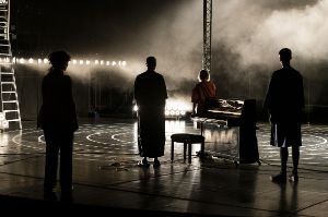 Four people stand backlit on a stage with their backs to the camera, a piano between them.
