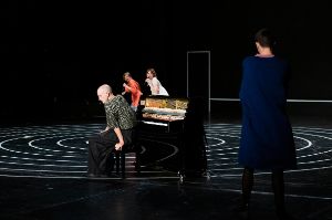 Two women sing, one man sits at a piano, another man watches.