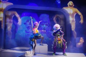 Eine Frau und ein Mann in bunten Kostümen stehen auf der Bühne, hinter ihnen ein großer Videoscreen mit animierten Fantasy-Figuren.