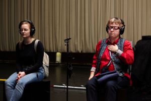 Two female read people sit next to each other. Both are wearing large headphones. The person on the right has visual physical impairments.