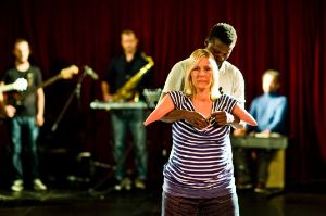 A female read person with arm deformity is held under the armpits by a male read person. He appears to be supporting her body weight. Behind them you can see a red stage curtain and three musicians with instruments.