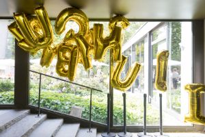 Goldene Buchstabenballons schweben unter der Decke, hinter ihnen ist ein großflächiges Fenster.