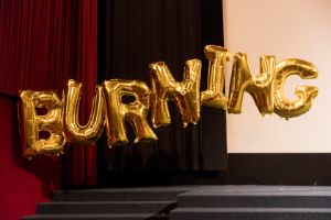 Golden letter balloons float in the air. Below and behind them is the stage. Left of them is a dark red stage curtain.