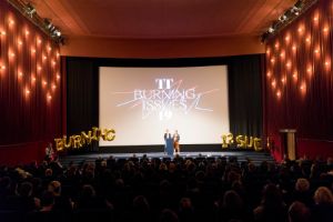 Frontal view of an illuminated stage. On the stage are two female readers. Behind them, "TT Burning Issues 2019" is projected on a screen. Flanking the stage are golden letter balloons forming the words "Burning Issues". Audience is seating in the auditorium.