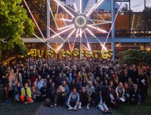 Group photo of the Burning Issues x Theatertreffen edition of 2019 in front of the glass facade of the Haus der Berliner Festspiele. The people in the holds golden letter balloons in the air forming the title "BURNING ISSUES".