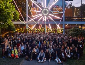 Gruppenfoto der Burning Issues x Theatertreffen-Ausgabe von 2019 vor der Glasfassade des Haus der Berliner Festspiele. Die hintere Menschenreihe hält goldene Buchstabenballons in die Luft, die den Titel „BURNING ISSUES“ bilden.