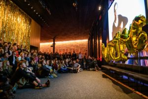 Many people sit in front of a screen and watch with a tense expression what is being shown. On the screen is a female read person. The setting on the screen reminds of an interview situation. Behind the audience is golden tinsel.