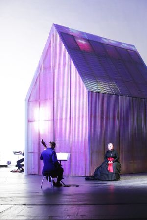 Das leicht pink leuchtende „Forecast House“ steht auf der Bühne, davor sitzen eine Frau in einem schwarzen Kleid und ein Musiker.