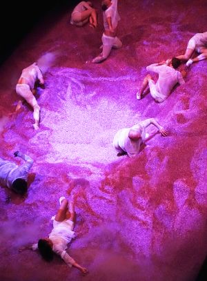 People move in mountains of pink granules on a stage.