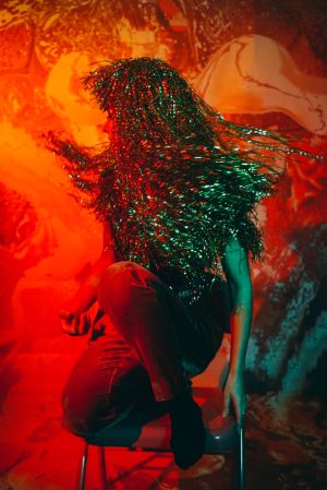 A person with a wig made of long tinsel threads squats on a chair. She swings energetically her head to the side.