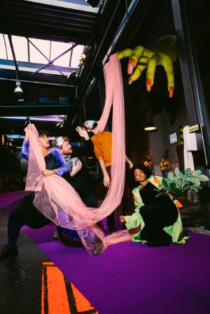 Four people pose with a tulle cape, a plant and with a disco ball. Above them is a set element representing a yellow grasping hand with long red nails