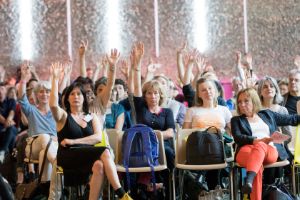 Frontal view of seated people in the audience. Most of them have their hand raised.