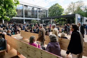 Many people are on the forecourt of the Haus der Berliner Festspiele in a labyrinth made of wooden benches.