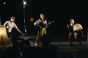 Three performers from "Bunker Cabaret" dance on stage, the person in the middle holds a guitar.