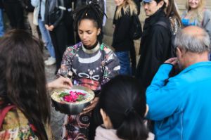 A person in a colourful dress holds a bowl with flower petals and green twigs, people around.