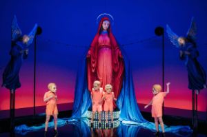 Four people in salmon pink costumes and with a mask over their heads stand in front of a huge statue of the Virgin Mary flanked by two angels.