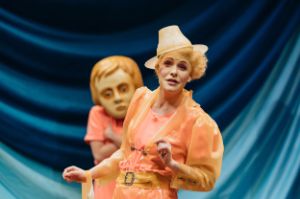 A woman with a slanted hat and her hair in curls stands in front of a person in a salmon-pink costume with a mask covering the head.