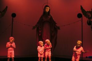 Four people in salmon-pink costumes with a mask over their heads stand in front of a huge statue of the Virgin Mary flanked by two angels.