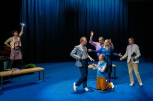 Six people in street clothes rehearse a scene on an empty stage.