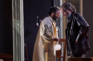 A younger man wearing a white coat and holding a test tube crosses an older man wearing a black leather jacket.