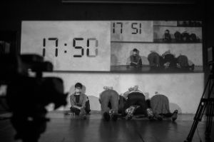 Three people are on a stage with their hands behind their backs, not facing the audience. Next to them sits a woman with a mask in front of her mouth. A clock on a screen shows 17:50.