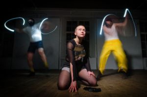 A woman crouches on the floor and looks up confidently. Behind her, two men dance with small lights in their hands.