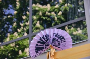 Ein lila Fächer mit dem Schriftzug „Theatertreffen“ befindet sich vor einem großen Fenster, in dem sich ein blühender Kastanienbaum spiegelt.