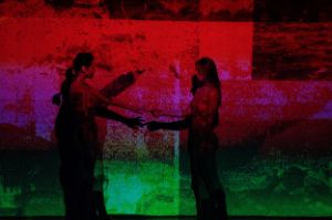 Two women join hands in front of a projection in red, purple and green.