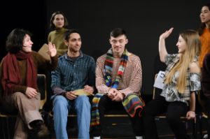 Two young women sit in a row of chairs and raise their hands to high five. Between them sit two smiling young men, behind them are two young women.