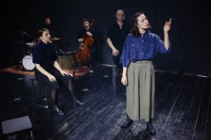 Eine Frau schaut auf ihre Hand, während sich hinter ihr Performer*innen und Musiker*innen befinden.