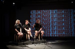 A woman and two men sit at microphone stands on a stage, with the title of the performance “PUTINPROCESS” projected on a screen in the background.