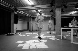 Two men and a woman in glittering costumes are in a performance space. Text sheets are spread on the floor, one of the men holds two of them in his hands.