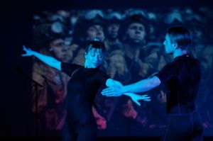 A woman and a man dance in front of a screen showing soldiers.