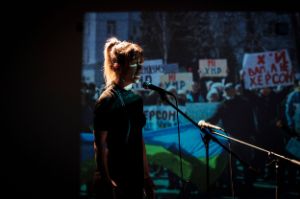 Eine Frau spricht vor einer Leinwand, die eine Demonstration zeigt.