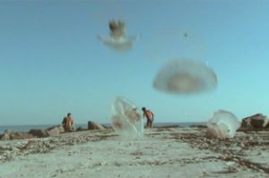 Jellyfish are falling on a beach, in the background are two fishermen with naked torsos.