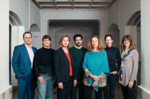 The seven Theatertreffen jury members stand in a white room and look into the camera.