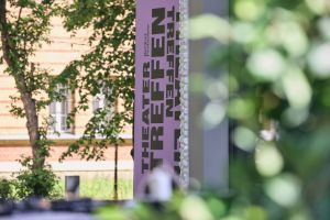 A Theatertreffen banner is amidst trees and buildings.